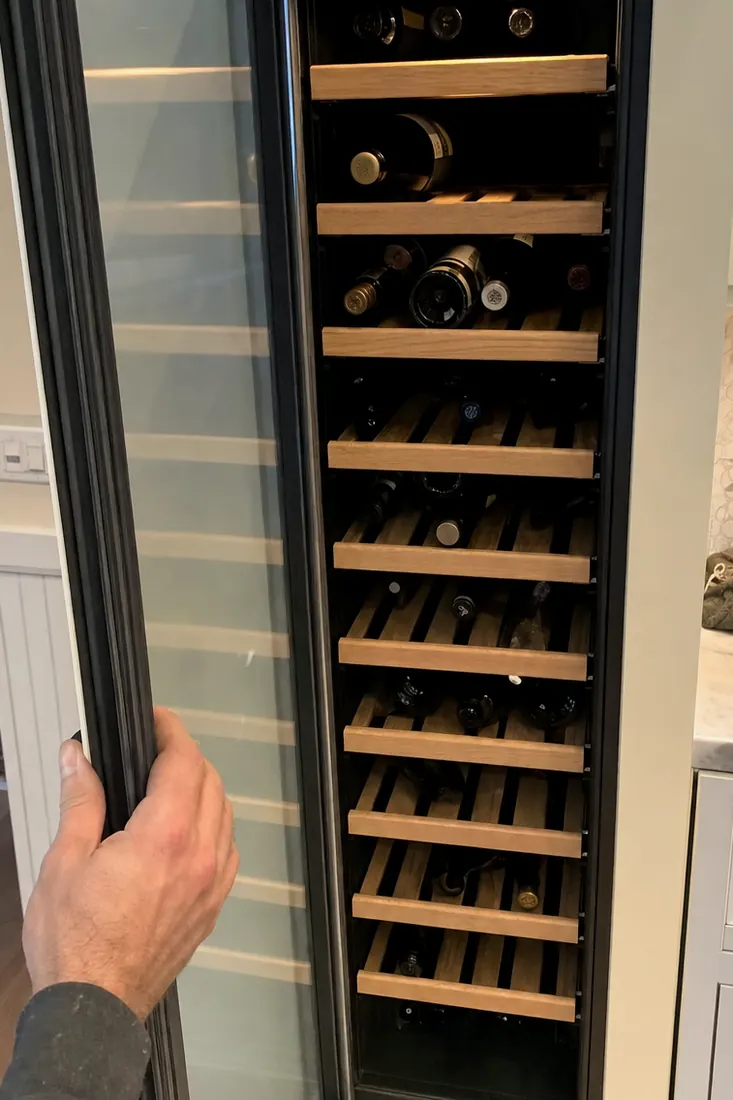 An open built-in wine column, bottles on wooden shelves, a hand at the door seal, the wooden shelves in frame - Sub-Zero and luxury appliance repair in Ponte Vedra Beach