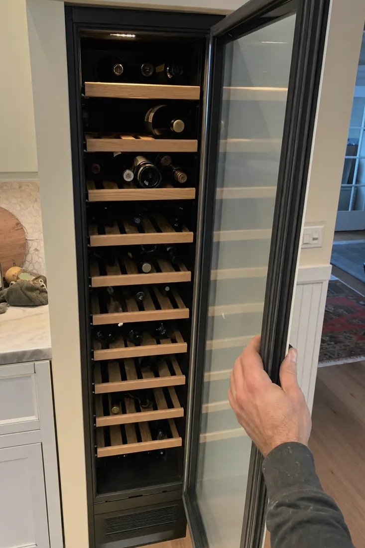 An open built-in wine column, bottles on wooden shelves, a hand at the door seal, the glass door in frame - Wine Cooler and Wine Storage Repair